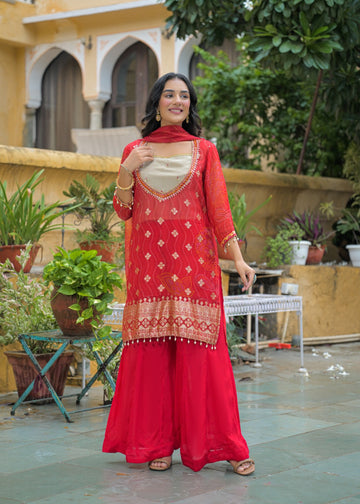 A woman stands outdoors wearing a bright red traditional outfit with a matching dupatta, featuring gold embroidery and a beige inner top. The setting includes potted plants, a stone patio, and a yellow building in the background.