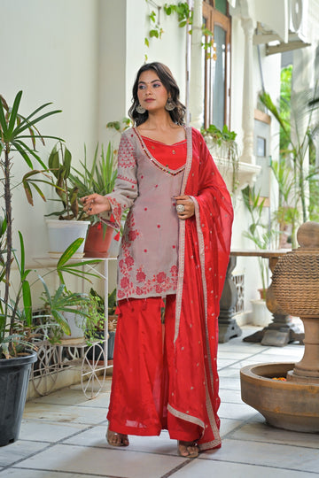 A woman models a traditional Indian salwar kameez suit set in a courtyard. The set features a light pinkish-beige kurta with red floral embroidery, a solid red inner lining and matching red pants, and a red dupatta with gold zari borders.