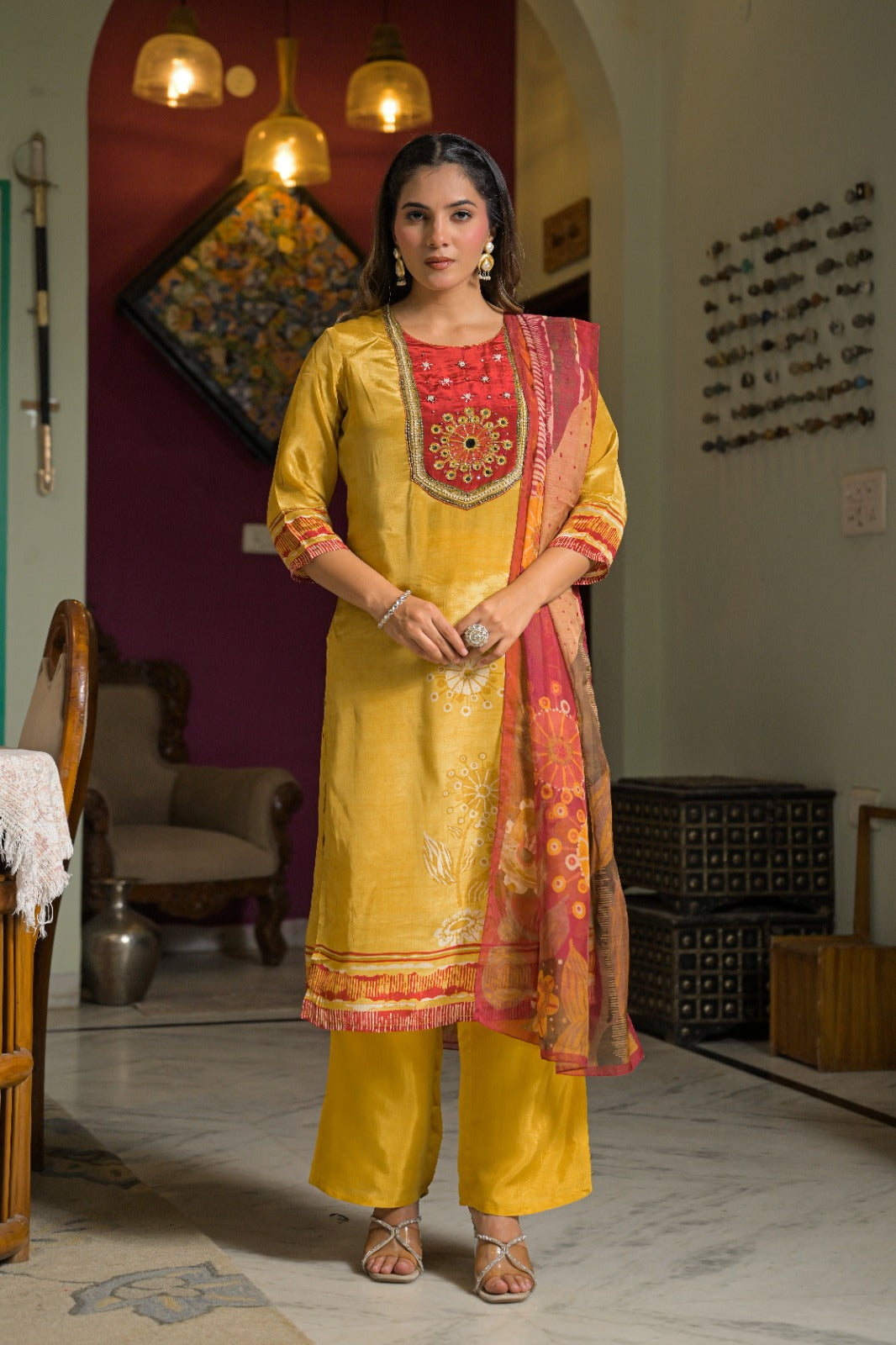 A woman stands indoors wearing a traditional Indian salwar kameez outfit in mustard yellow, featuring a red embroidered yoke, matching trousers, and a pink and gold dupatta.