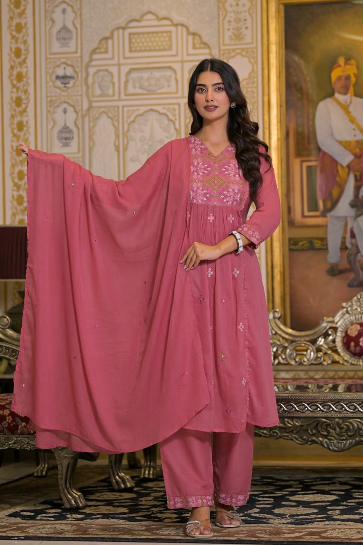 A woman models a pink traditional Indian salwar kameez suit with a matching dupatta in an ornate indoor setting. The outfit features a pink tunic with floral embroidery and matching trousers.