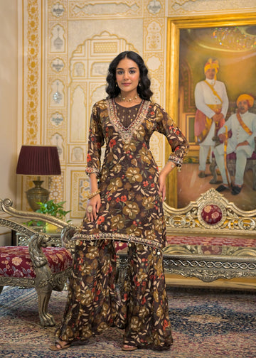 A woman models a brown three-piece printed kurta set with floral motifs and gold embroidery, standing in an ornate traditional room with a carved sofa and a large framed painting.