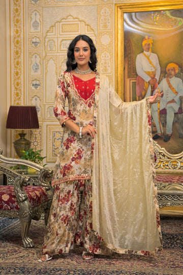 A woman models a traditional South Asian salwar kameez suit in an ornate, vintage-style room. The outfit features a beige and red floral print kameez with a solid red embroidered neckline and a matching beige dupatta.