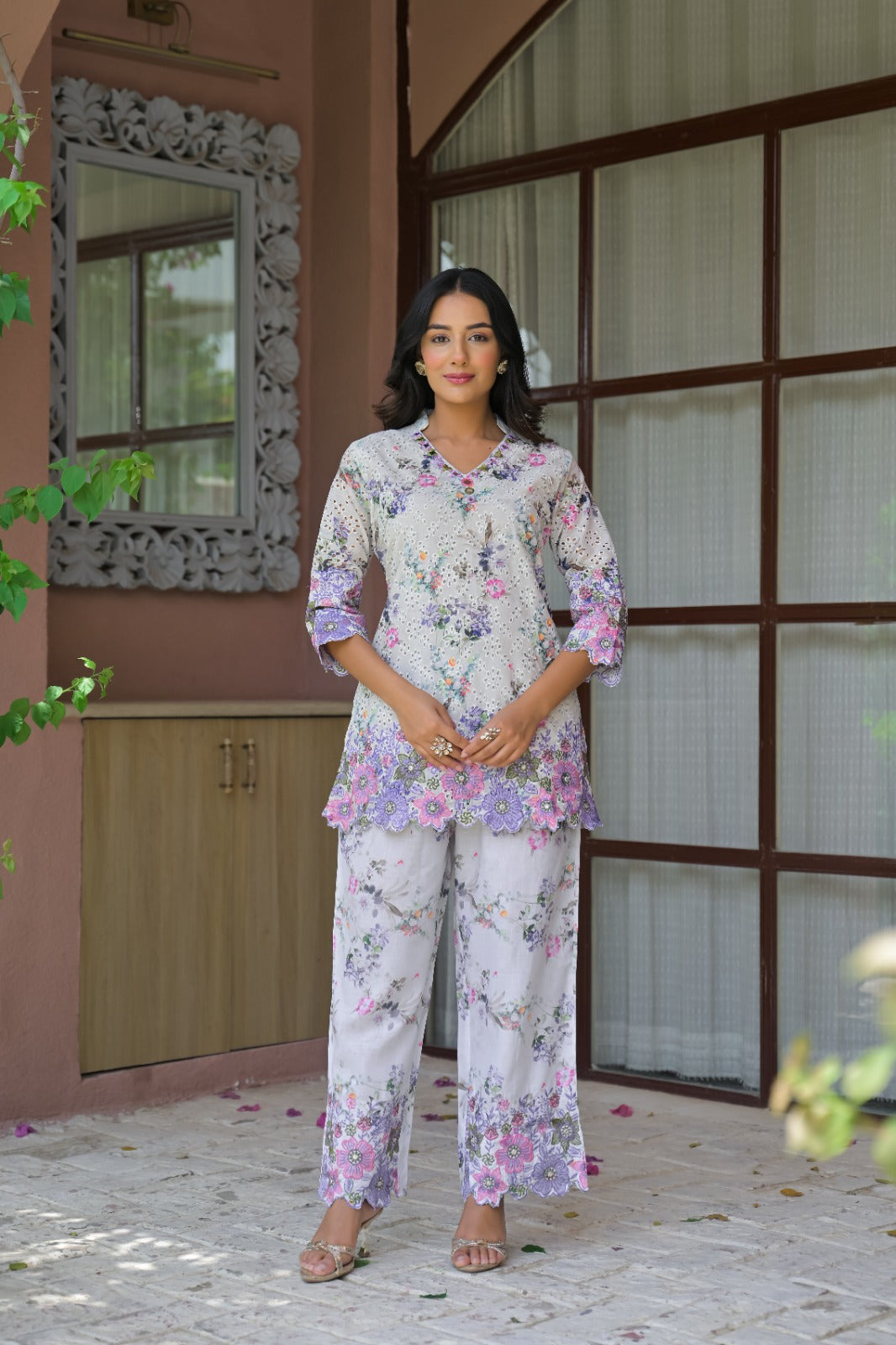 A woman modeling a white two-piece floral pajama set with pink and purple accents, standing in front of a rustic brown door and pink wall.
