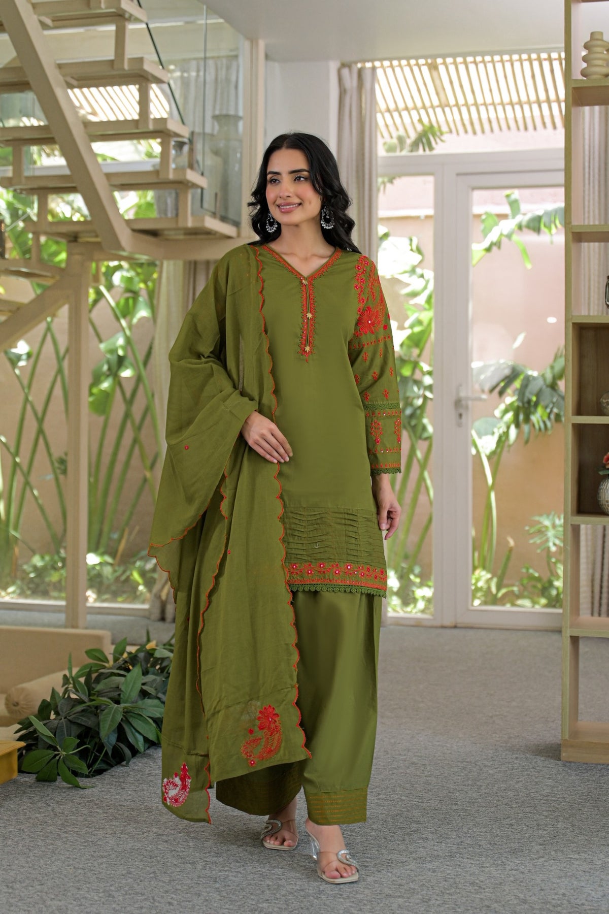 A woman models an olive green three-piece salwar kameez suit with red embroidered details on the kurta and dupatta, set against a modern indoor background with a staircase and glass doors.