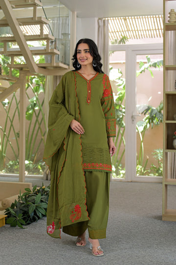 A woman models an olive green three-piece salwar kameez suit with red embroidered details on the kurta and dupatta, set against a modern indoor background with a staircase and glass doors.
