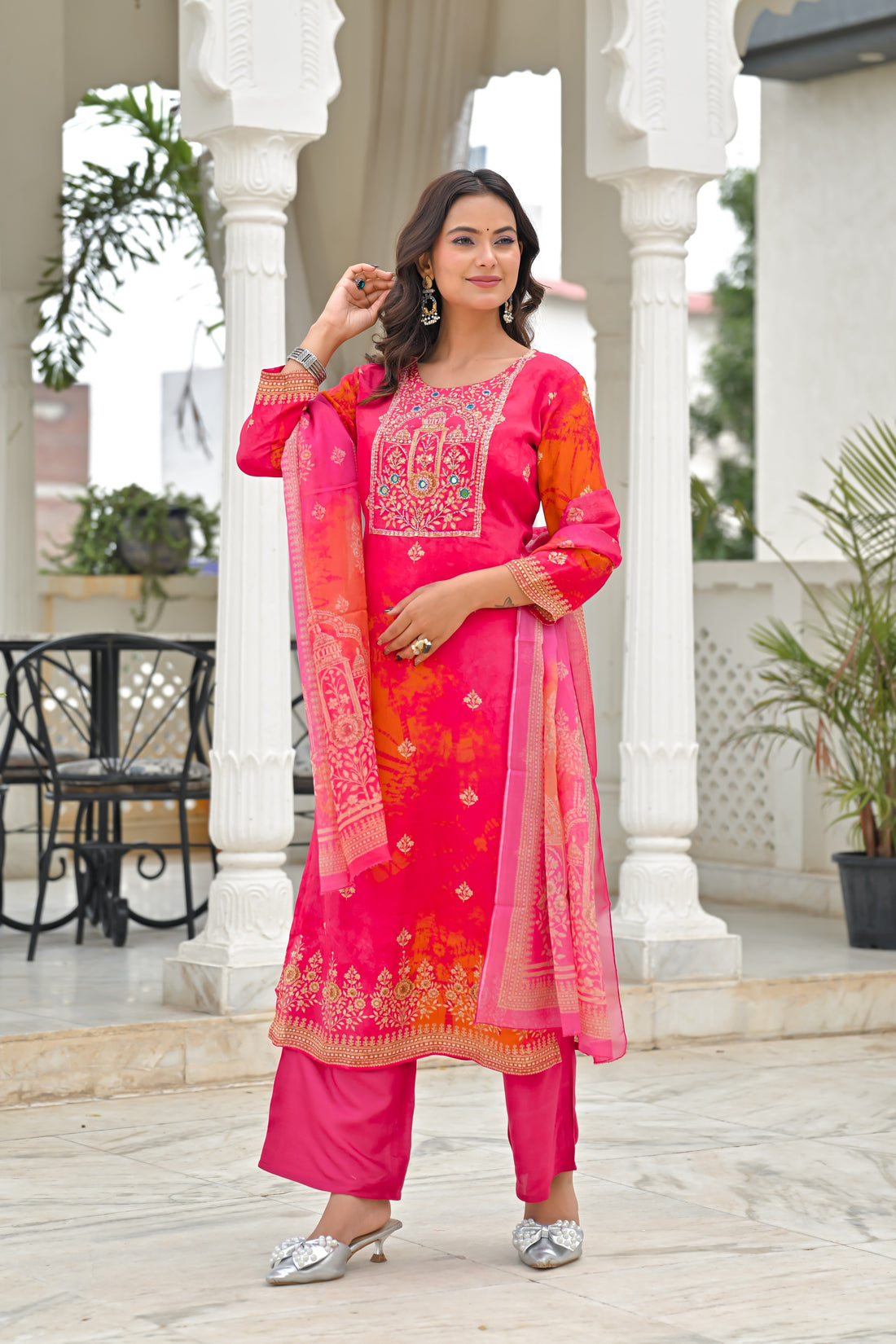 A woman models a bright pink Indian ethnic salwar kameez suit with matching trousers and a dupatta, featuring orange and yellow floral patterns and gold embroidery, set against a white architectural background.