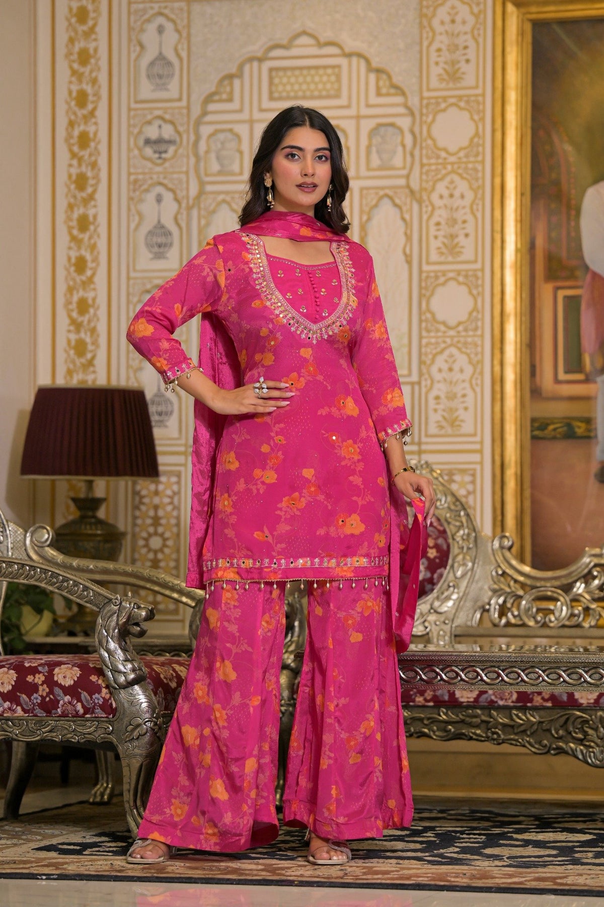 A woman models a bright pink traditional Indian salwar kameez suit with yellow floral patterns and gold embroidery, standing in an ornate, vintage-style room with gold-trimmed furniture.