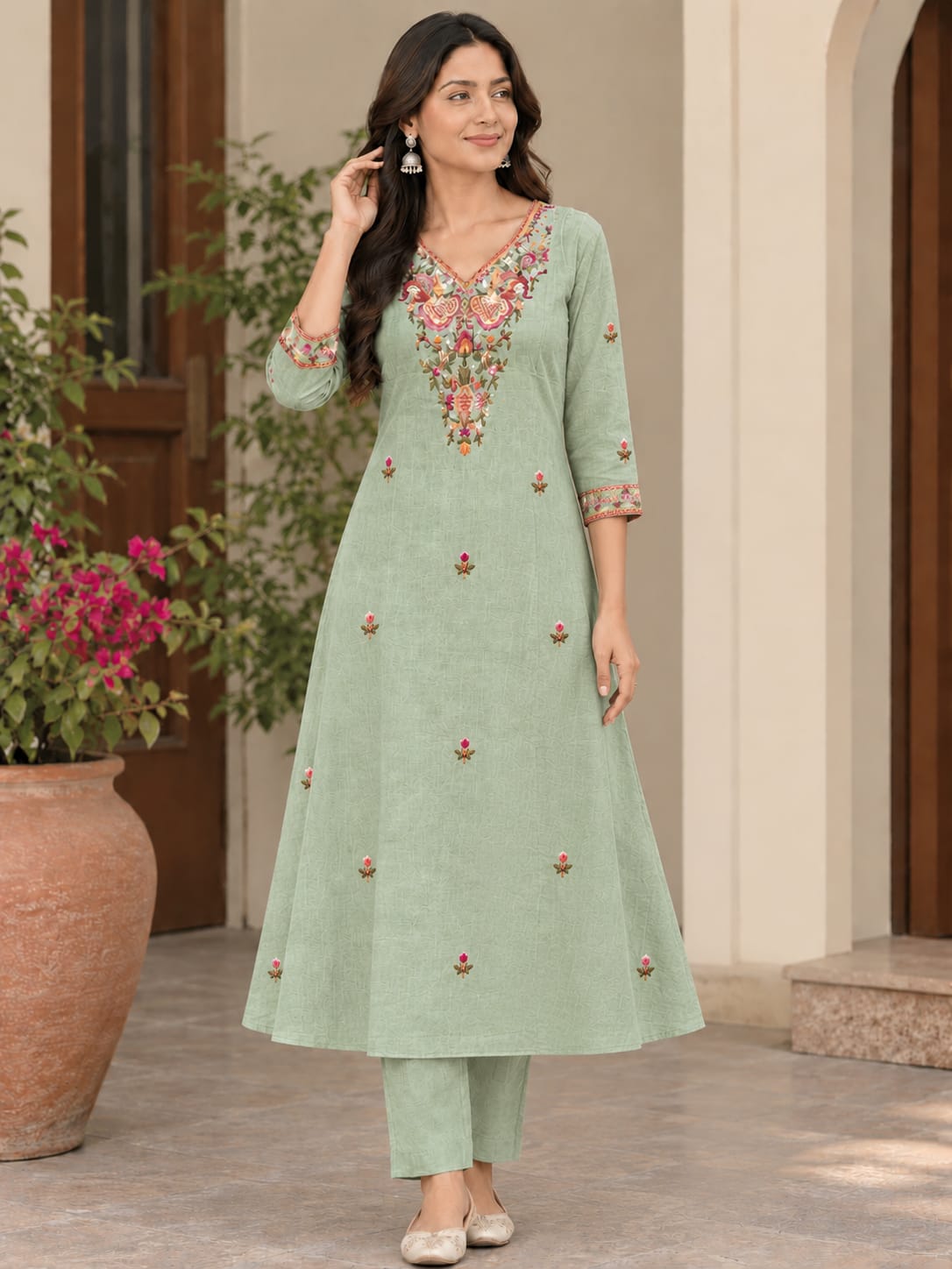 A woman models a light sage green A-line kurta with colorful floral embroidery on the neckline and scattered embroidered motifs on the front, paired with matching green trousers in an outdoor courtyard setting.