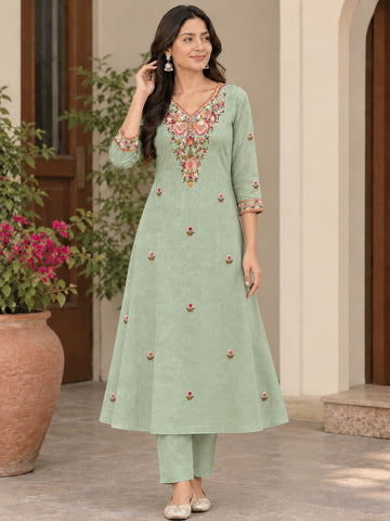 A woman models a light sage green A-line kurta with colorful floral embroidery on the neckline and scattered embroidered motifs on the front, paired with matching green trousers in an outdoor courtyard setting.