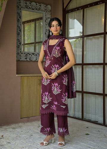 A woman models a three-piece salwar kameez suit in deep purple with pink and white floral embroidery, standing in front of a brown wooden window and wall.