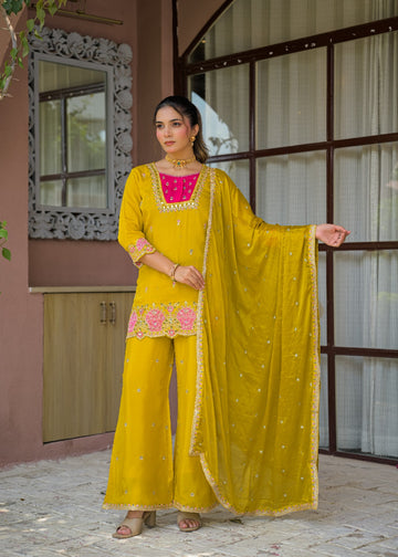A woman models a traditional Indian salwar kameez suit in mustard yellow, featuring a bright pink embroidered yoke, matching wide-leg pants, and a sheer embroidered dupatta, set against a rustic building facade.