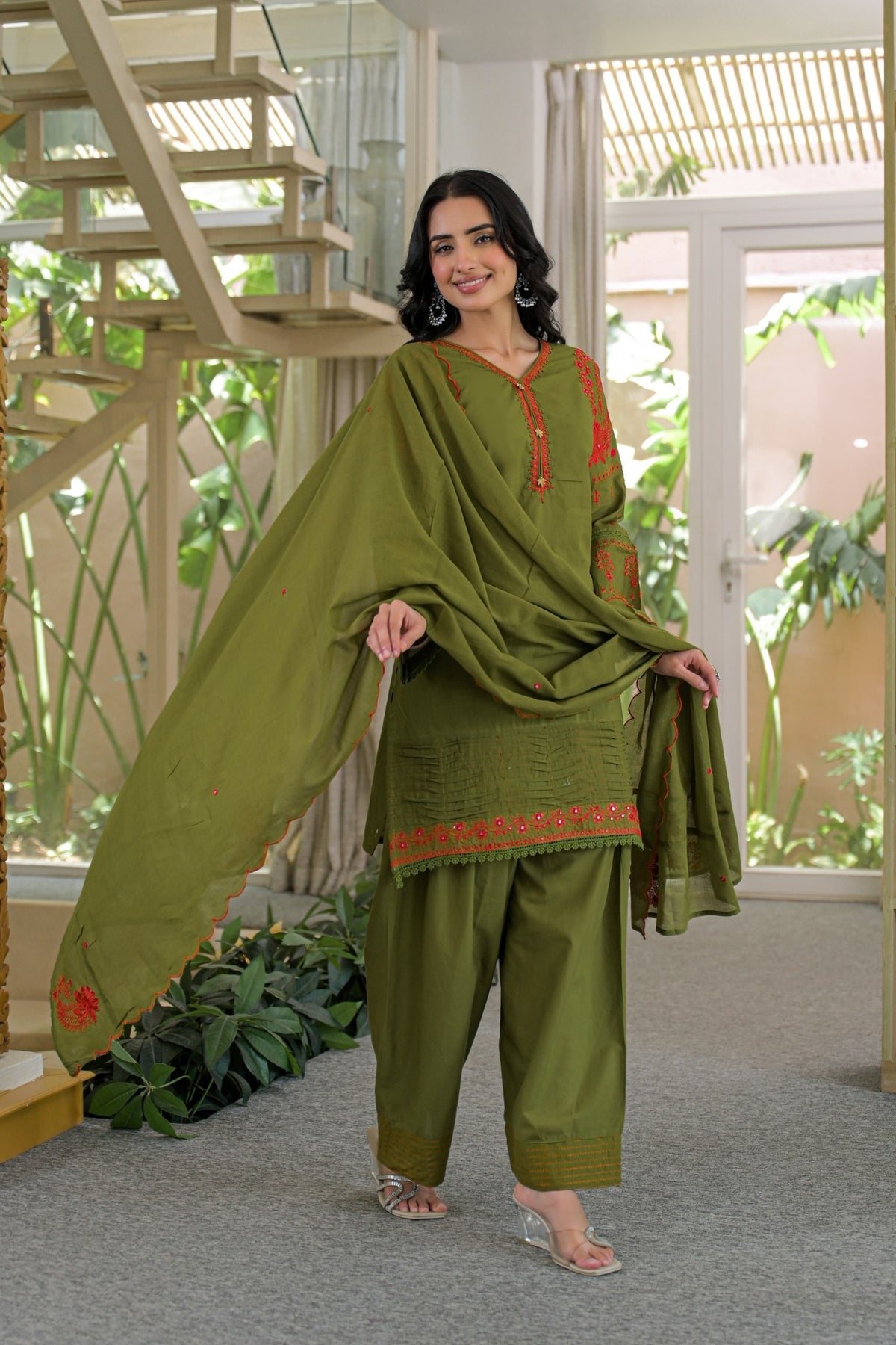 A woman models an olive green ethnic three-piece suit set with red embroidered details, featuring a kurta, wide-leg pants, and a matching dupatta, set against an indoor background with a staircase and plants.