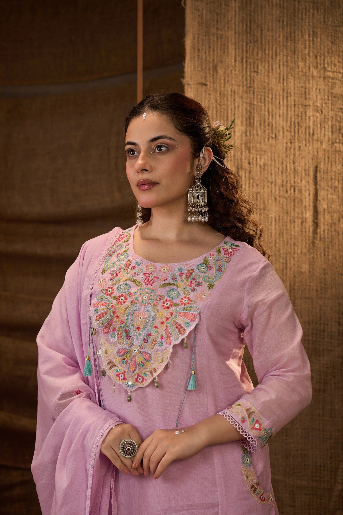 A woman stands in a rustic indoor setting wearing a light pink traditional outfit with colorful embroidered details on the chest and lower kurta, paired with a matching dupatta. The background features a burlap curtain and stacked terracotta pots.