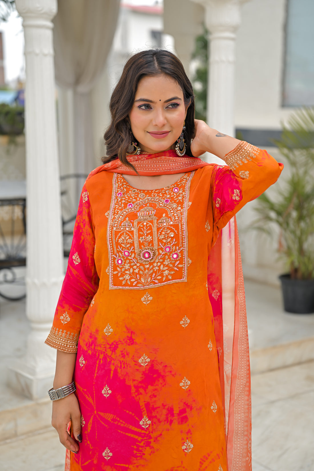 A woman models a bright pink Indian ethnic salwar kameez suit with matching trousers and a dupatta, featuring orange and yellow floral patterns and gold embroidery, set against a white architectural background.