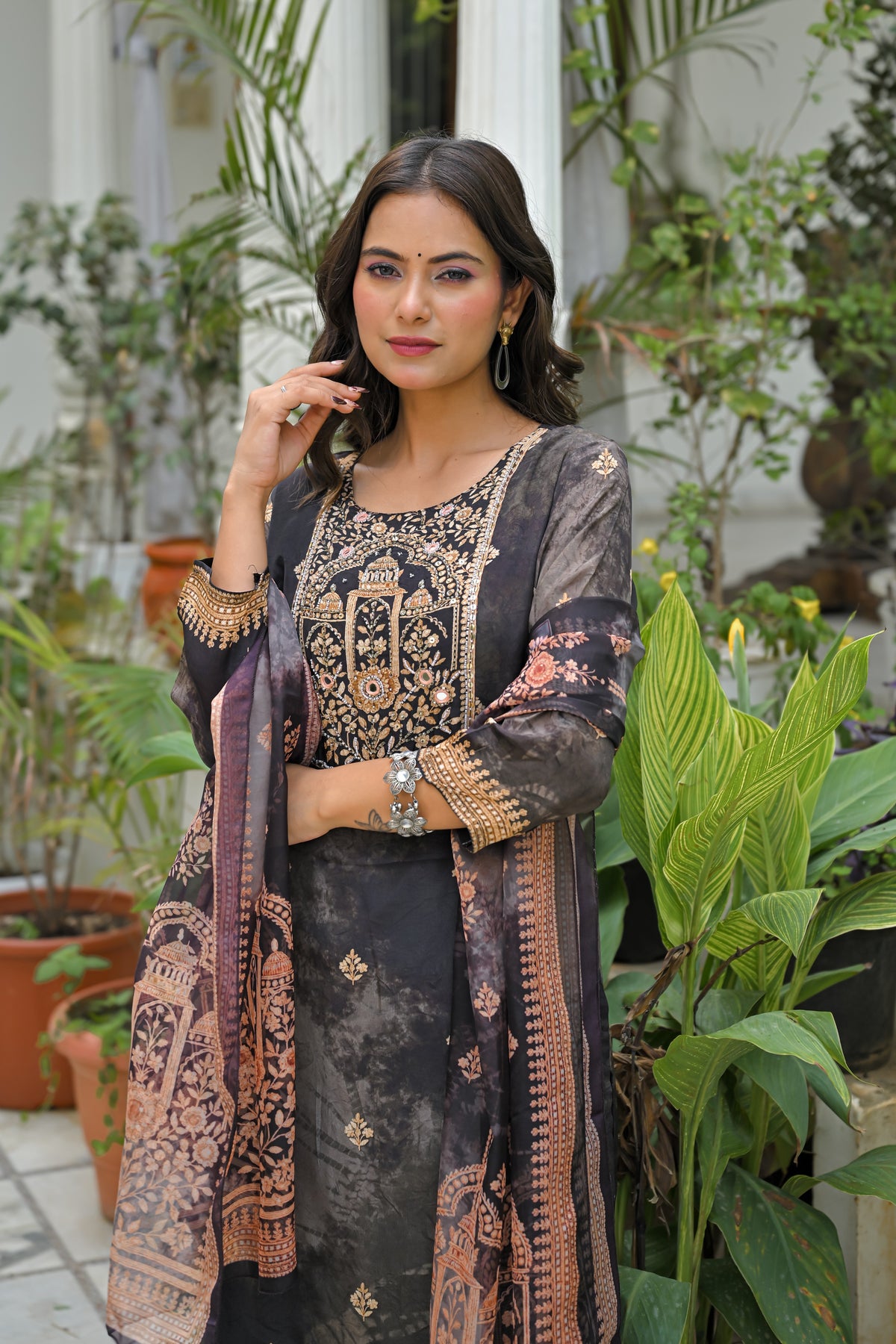 A woman models a traditional Indian salwar kameez suit in a garden setting. The outfit features a black base with intricate gold embroidery on the chest and sleeves, paired with a matching patterned dupatta.