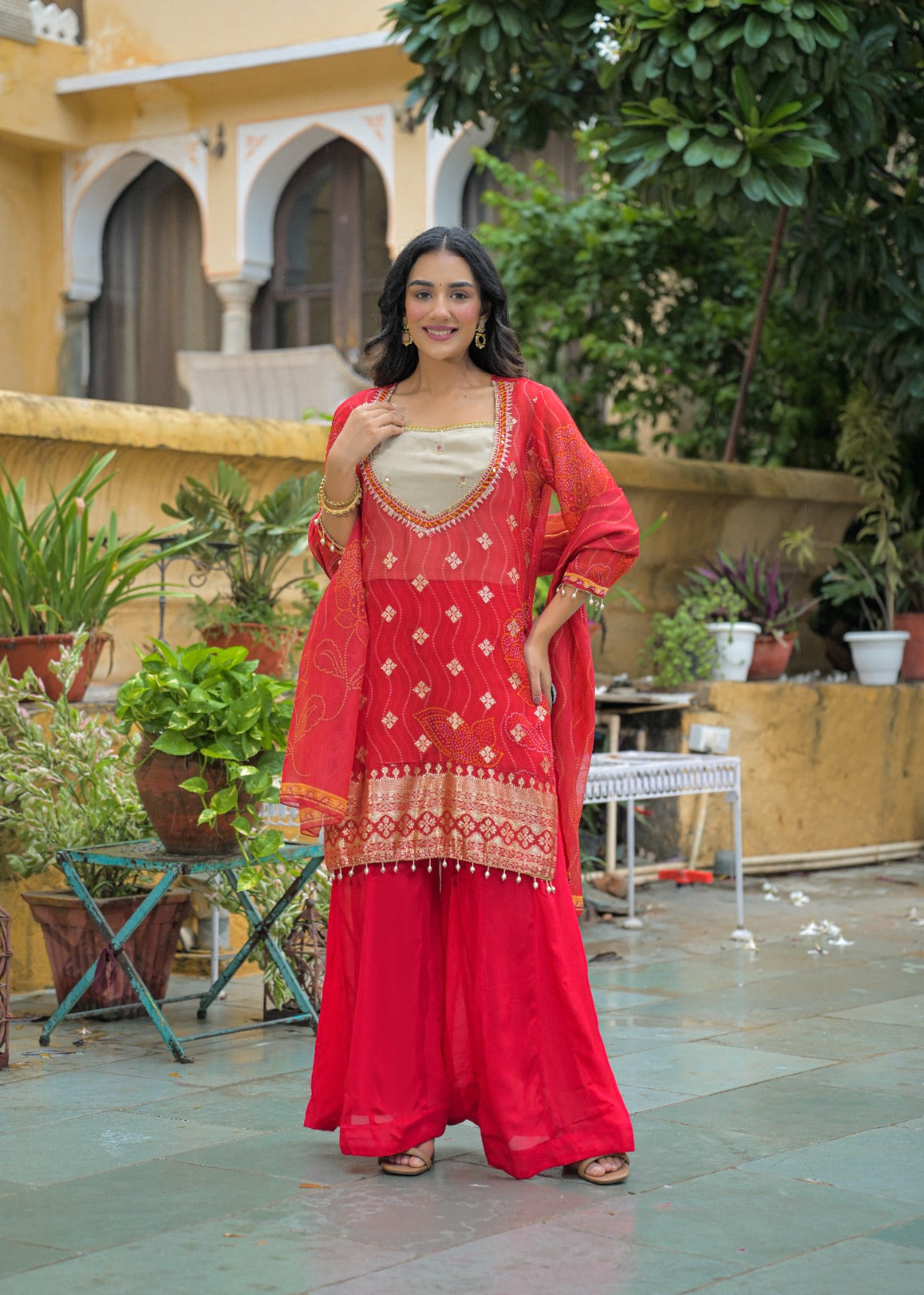 A woman models a traditional Indian red salwar kameez suit with gold embroidery, a cream inner kurta, and a red dupatta in an outdoor courtyard setting.