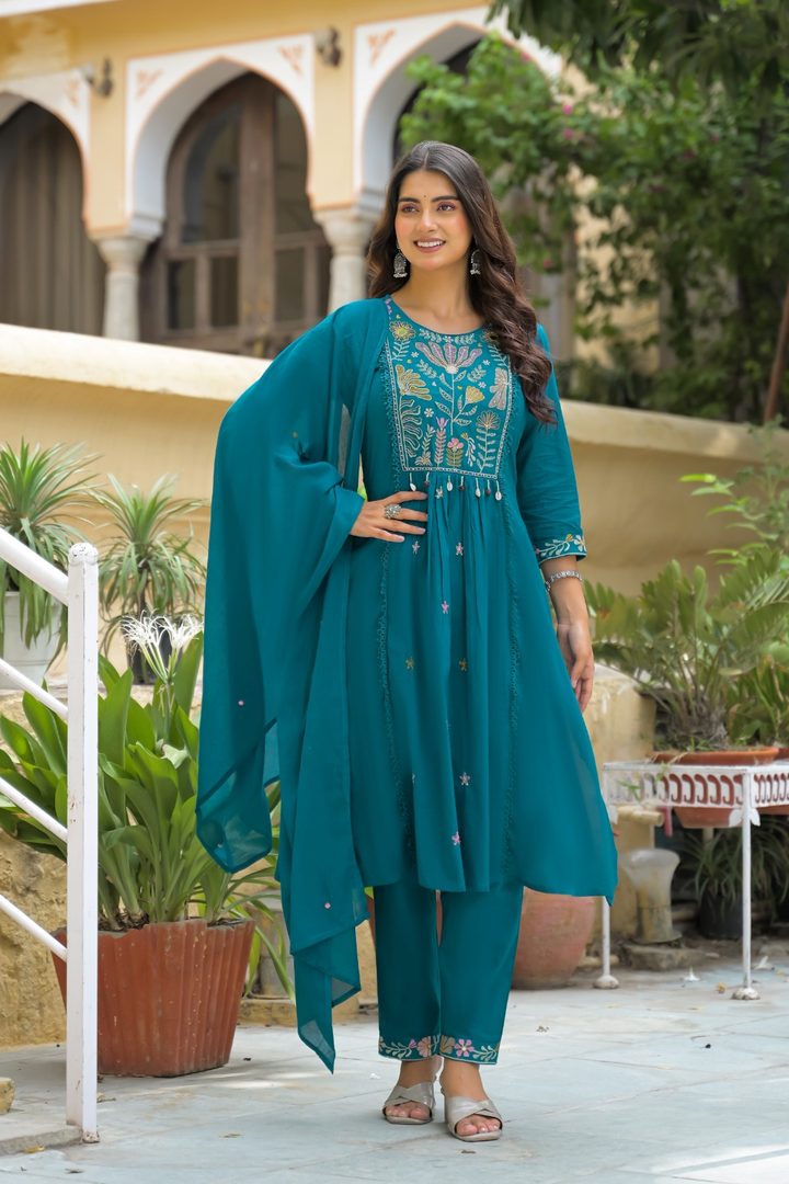 A woman models a teal blue ethnic salwar kameez suit with intricate embroidery, featuring a matching dupatta, in an outdoor courtyard setting.