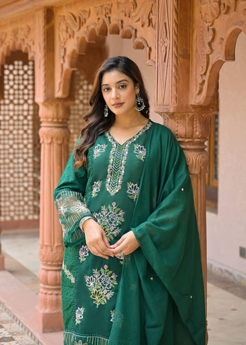 A woman wearing a green embroidered traditional outfit with a matching dupatta, standing in front of ornate carved stone pillars.