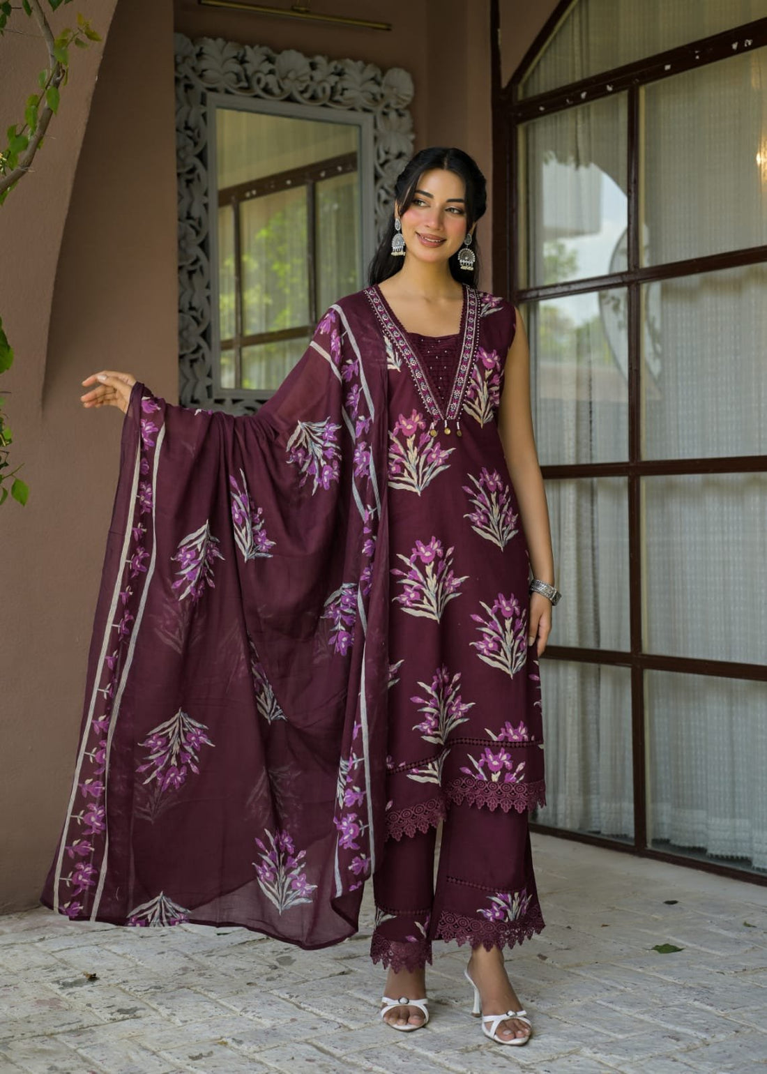 A woman models a three-piece salwar kameez suit in deep purple with pink and white floral embroidery, standing in front of a brown wooden window and wall.