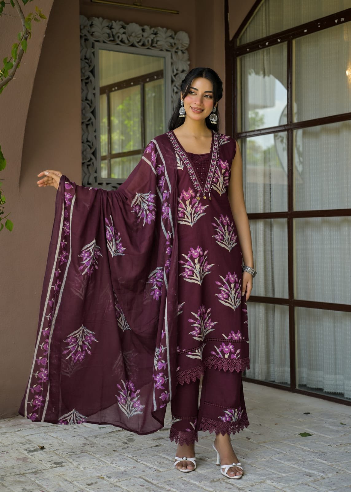 A woman models a three-piece maroon ethnic salwar kameez suit with floral embroidery, standing in front of a building with large window panes.