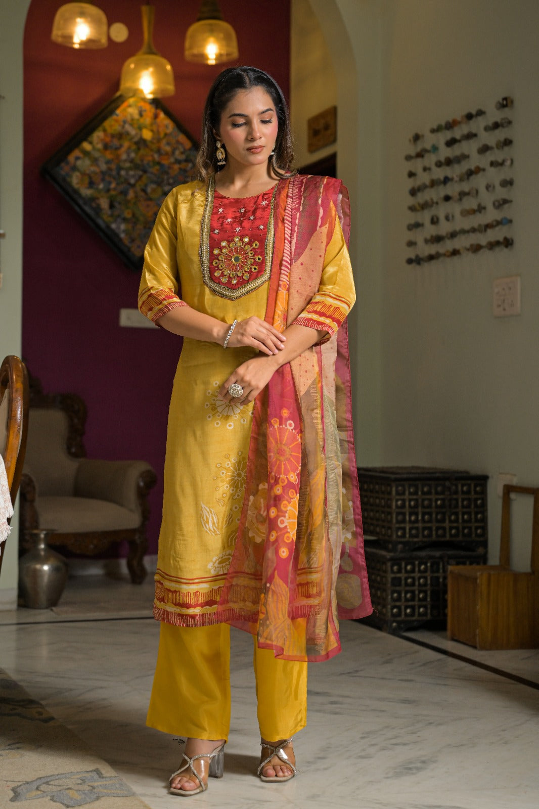 A woman stands indoors wearing a traditional Indian salwar kameez outfit in mustard yellow, featuring a red embroidered yoke, matching trousers, and a pink and gold dupatta.