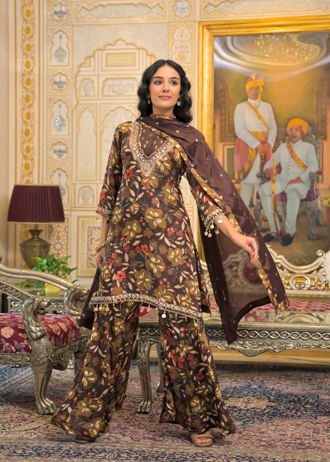 A woman models a brown floral printed salwar kameez suit with matching wide-leg pants and a sheer dupatta in an ornate, traditional Indian room with gold accents.