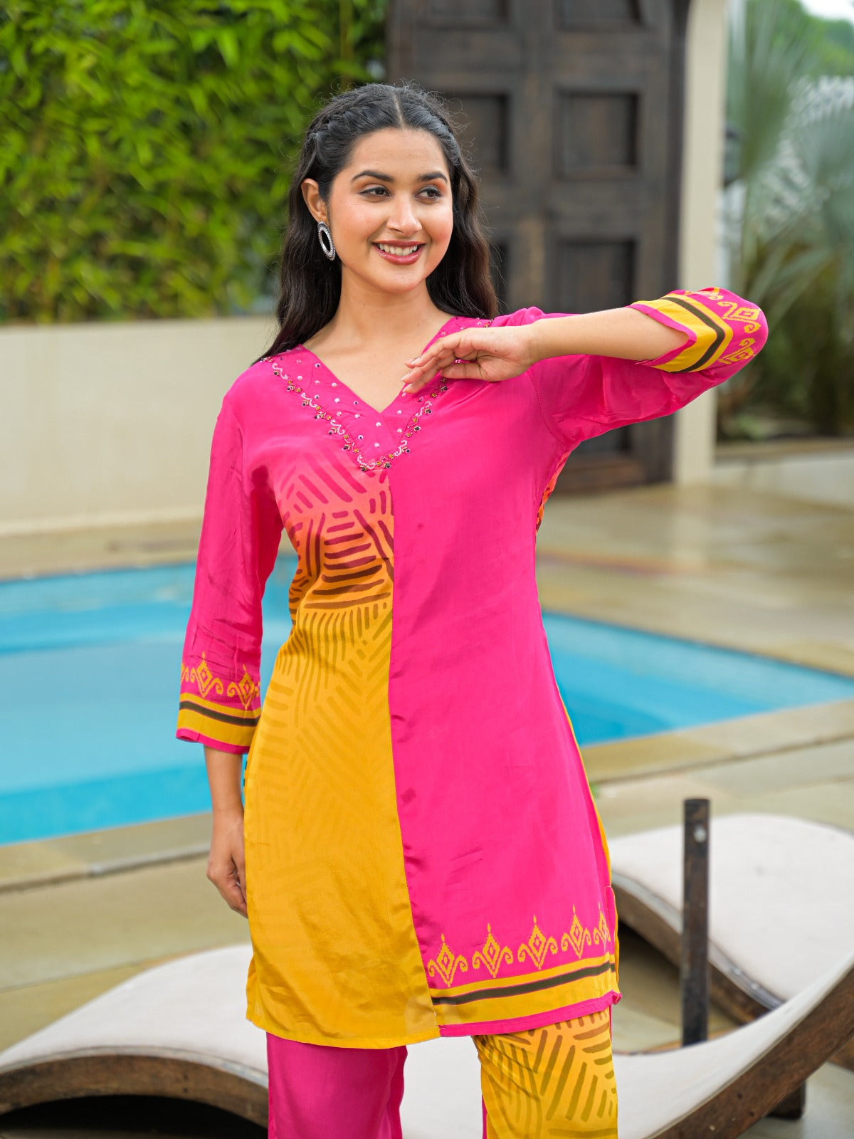 A woman modeling a bright pink and yellow color-blocked kurta set with matching pants, standing outdoors next to a swimming pool.