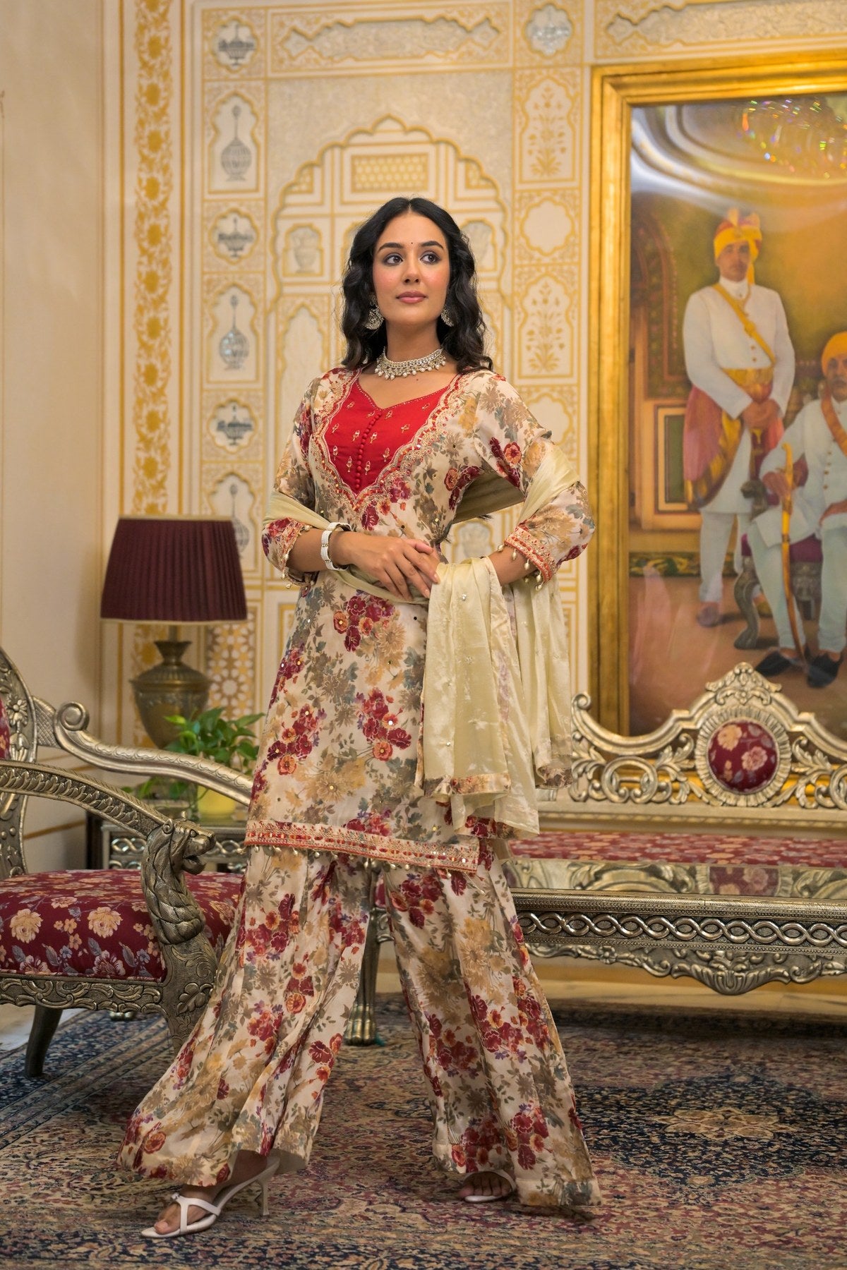 A woman models a traditional Indian salwar kameez suit in an ornate, vintage-style room. The outfit features a beige and red floral print with a solid red inner bodice (kurta) and a cream dupatta.