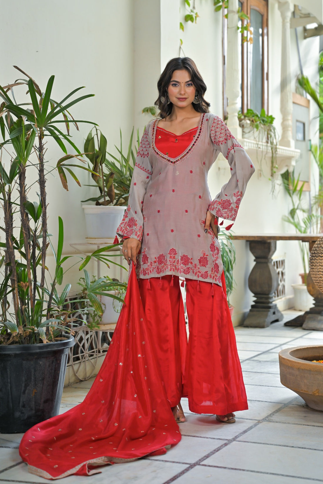 A woman models a traditional Indian salwar kameez suit set in a courtyard. The set features a light pinkish-beige kurta with red floral embroidery, a solid red inner lining and matching red pants, and a red dupatta with gold zari borders.