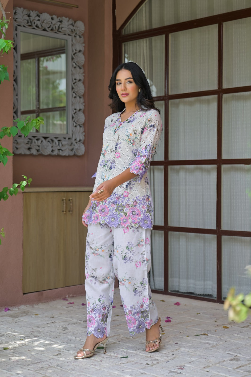A woman models a two-piece floral pajama set featuring a matching tunic top and wide-leg pants, set against an outdoor patio backdrop with a pink wall and window frames.