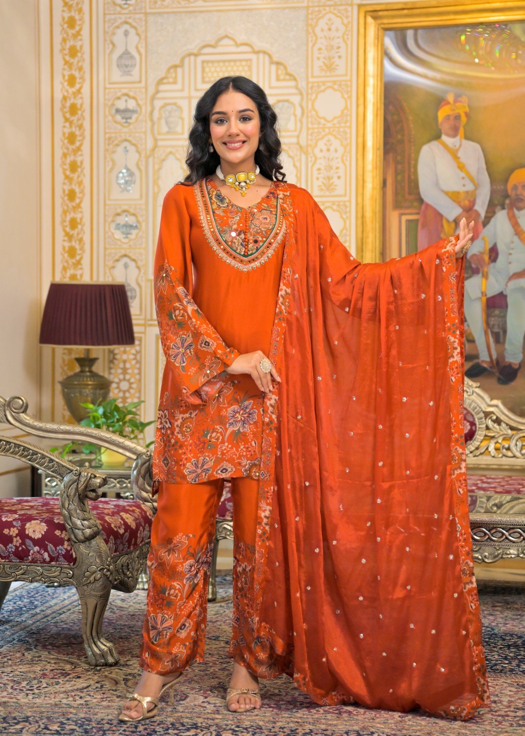 A woman models a traditional Indian salwar kameez suit in a rich orange color with intricate gold and silver embroidery, paired with a matching sheer dupatta. The setting is an ornate, vintage-style room with gold-trimmed walls, a large painting, and antique furniture.