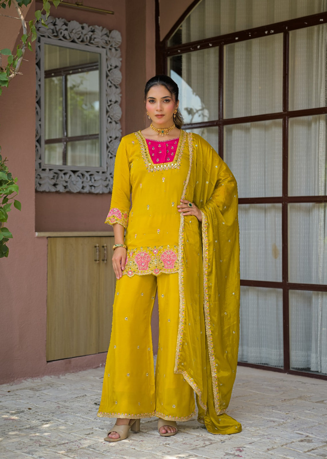A woman models a traditional Indian salwar kameez suit in mustard yellow, featuring a bright pink embroidered yoke, matching wide-leg pants, and a sheer embroidered dupatta, set against a rustic building facade.