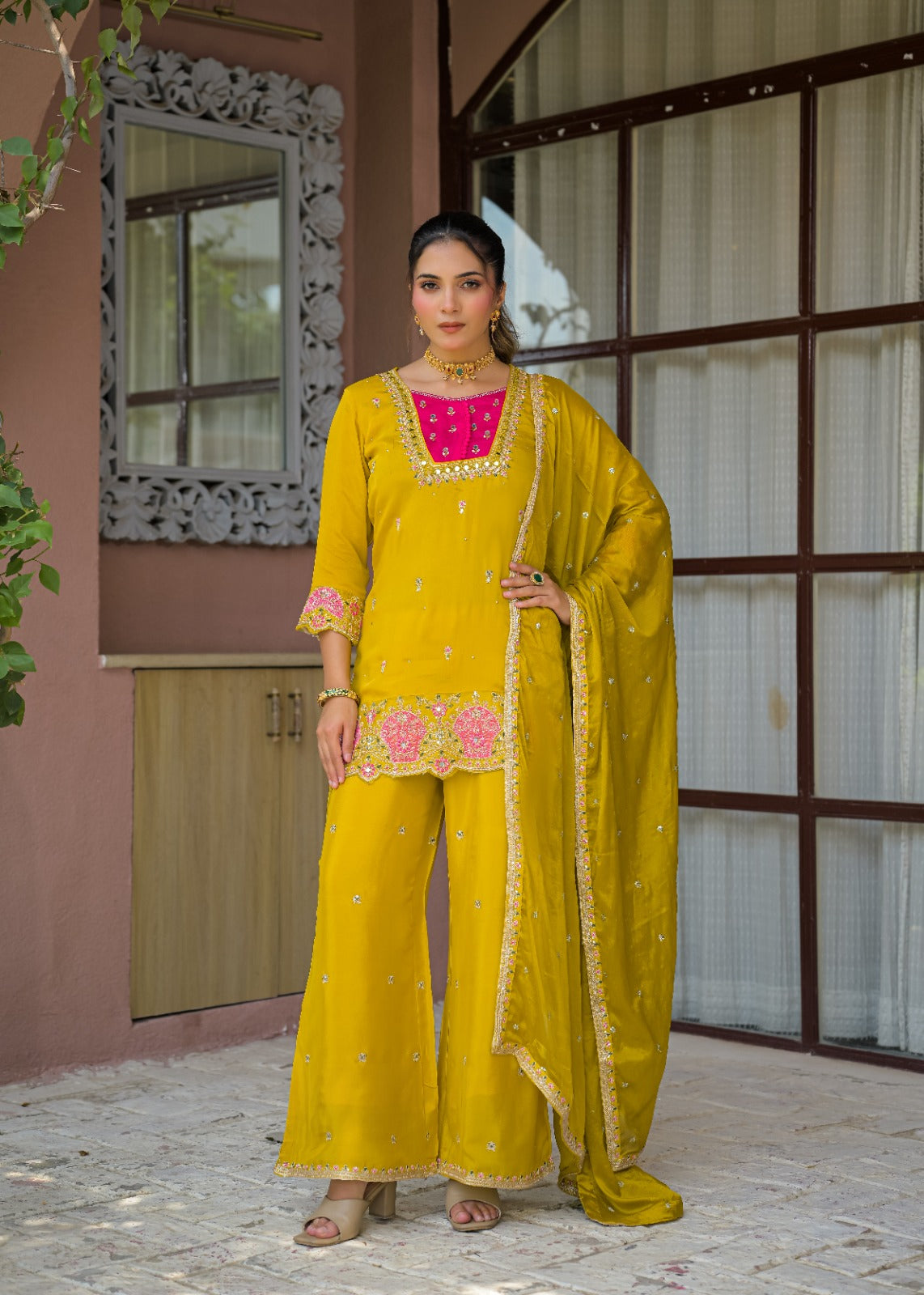 A woman models a traditional Indian salwar kameez suit in mustard yellow with pink and gold embroidery, standing in front of a building with a pink wall and window frames.
