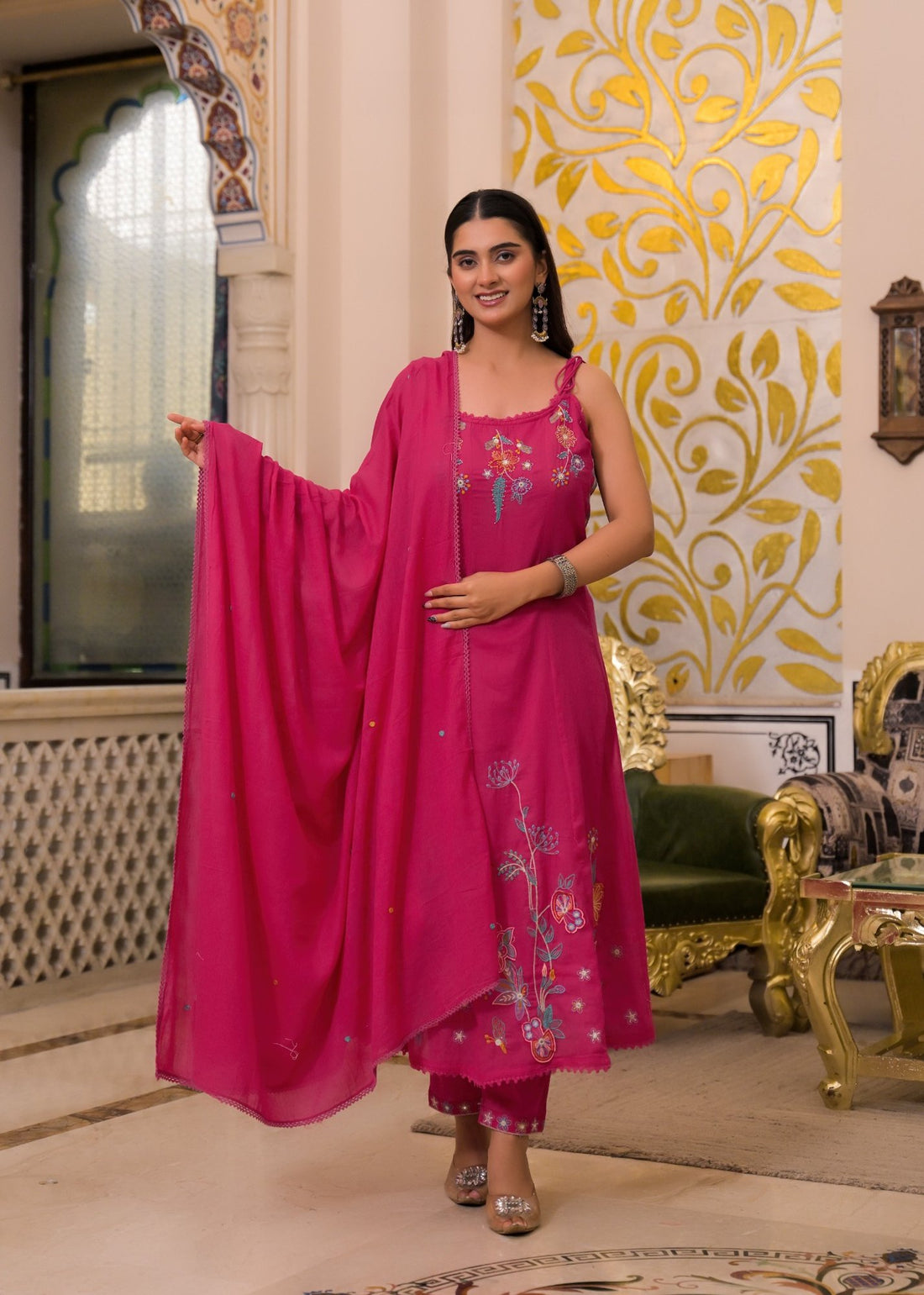 A woman modeling a bright pink embroidered kurta set with a dupatta in an ornate indoor setting with gold-patterned walls and green-gold furniture.