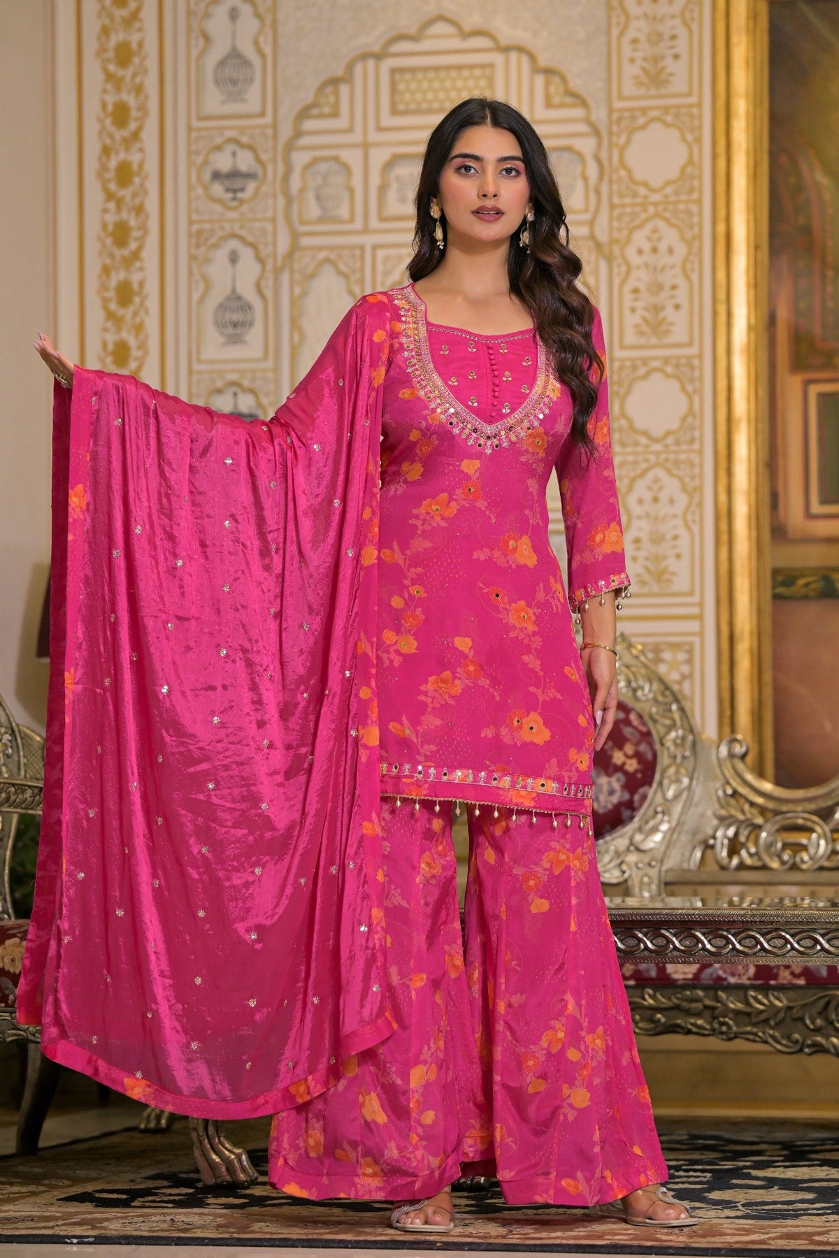 A woman models a bright pink traditional South Asian salwar kameez suit with a matching dupatta, featuring gold floral patterns and silver mirror work, set against an ornate golden interior background.