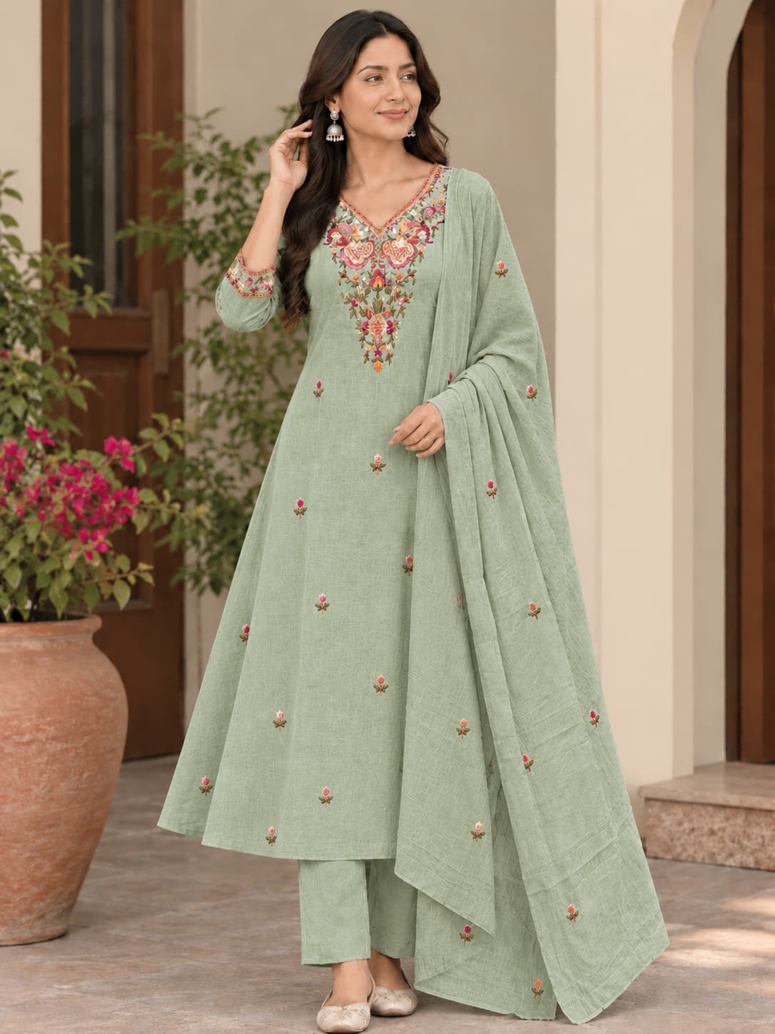 A woman models a sage green salwar kameez suit with floral embroidery on the neckline and scattered embroidered details on the fabric, paired with matching trousers and a dupatta, set against an outdoor architectural background with a potted plant.