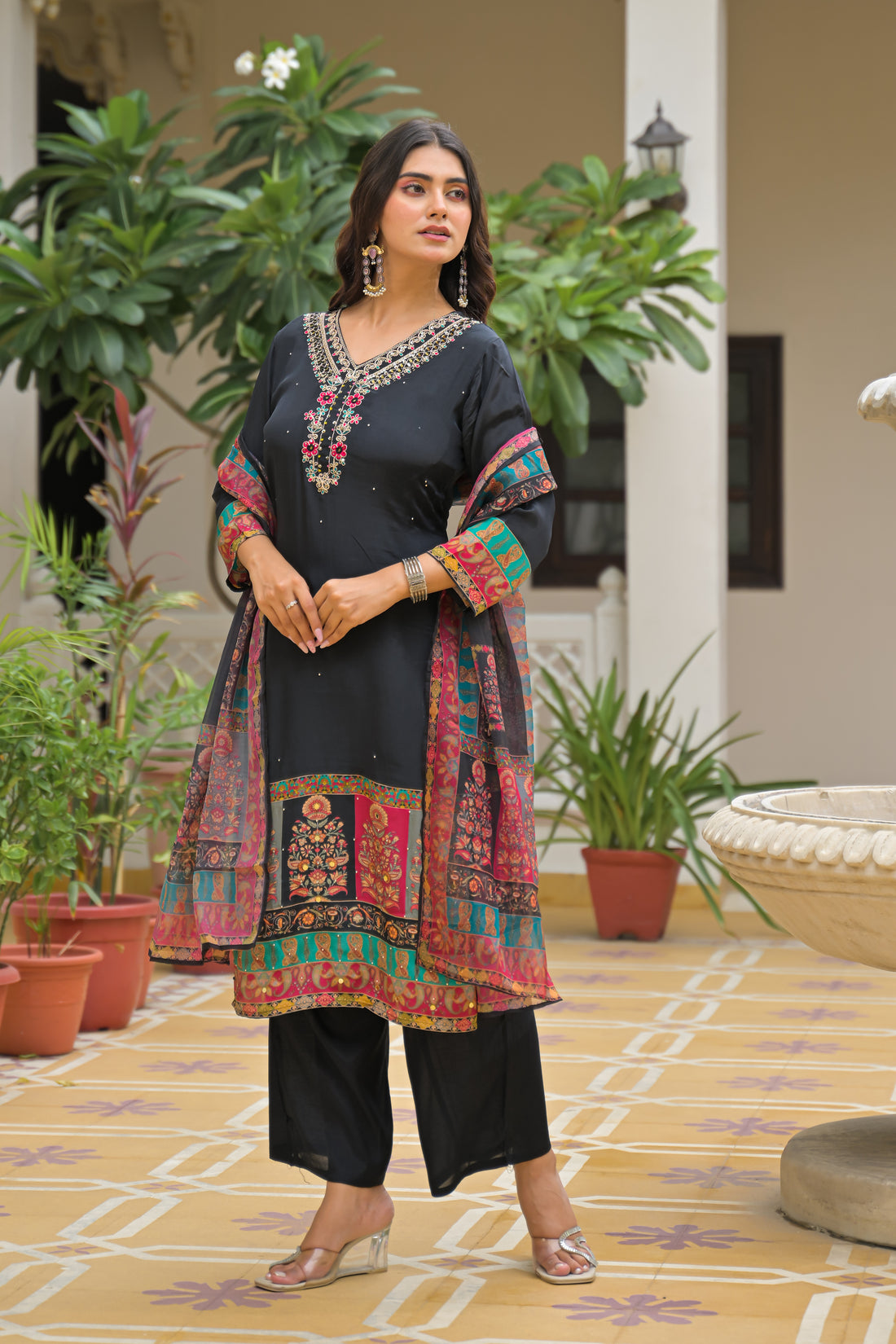 A woman models a black three-piece salwar kameez suit with colorful printed borders and a matching dupatta in an outdoor courtyard setting.