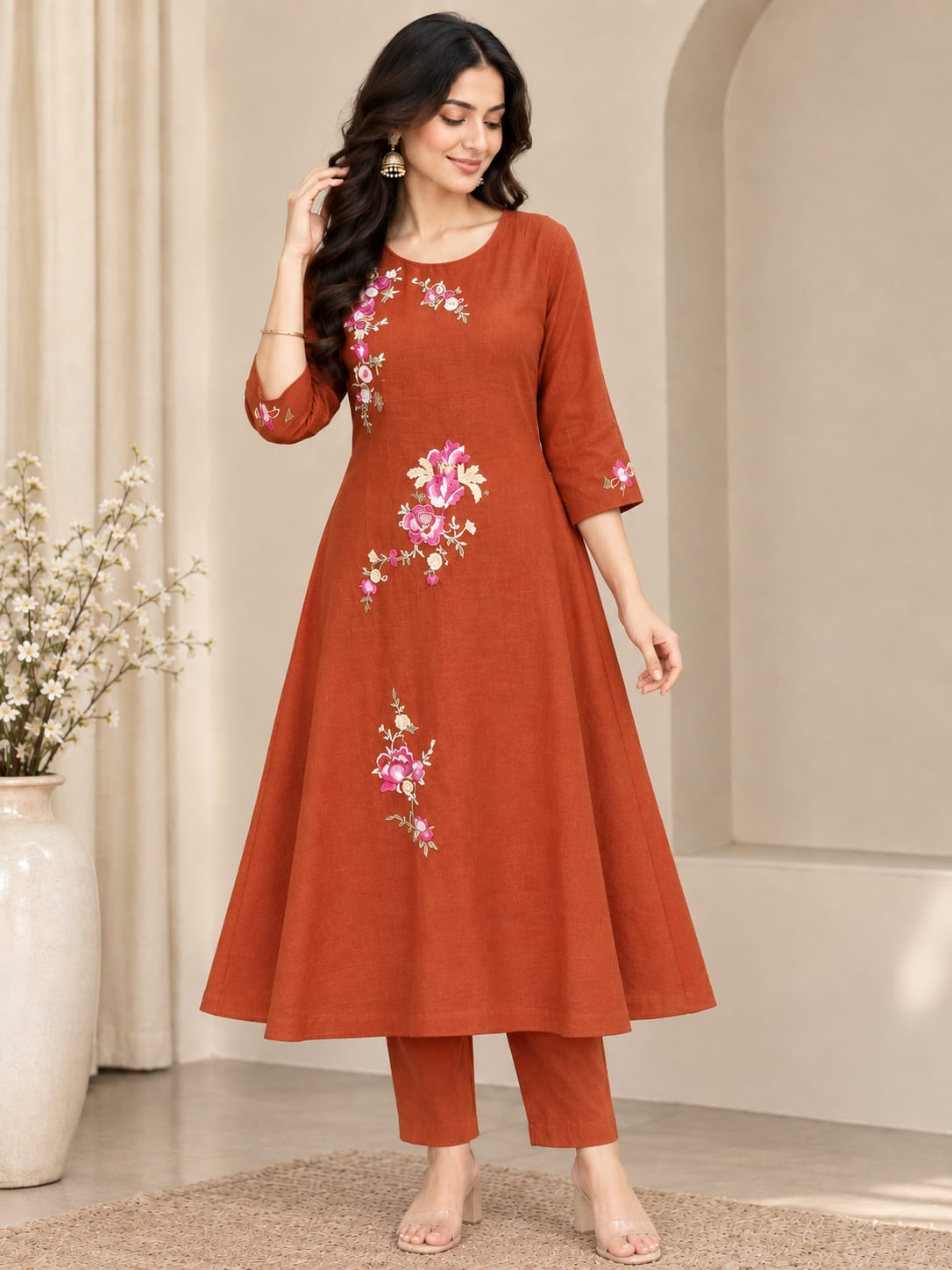 A woman models a rust-orange three-piece salwar kameez suit set with floral embroidery, featuring a long kurta, matching trousers, and a draped dupatta, set against a neutral indoor background.