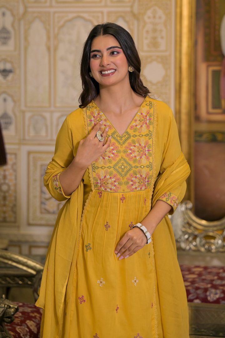 A woman wearing a yellow embroidered kurta with a matching dupatta, standing in an ornate indoor setting with patterned walls.