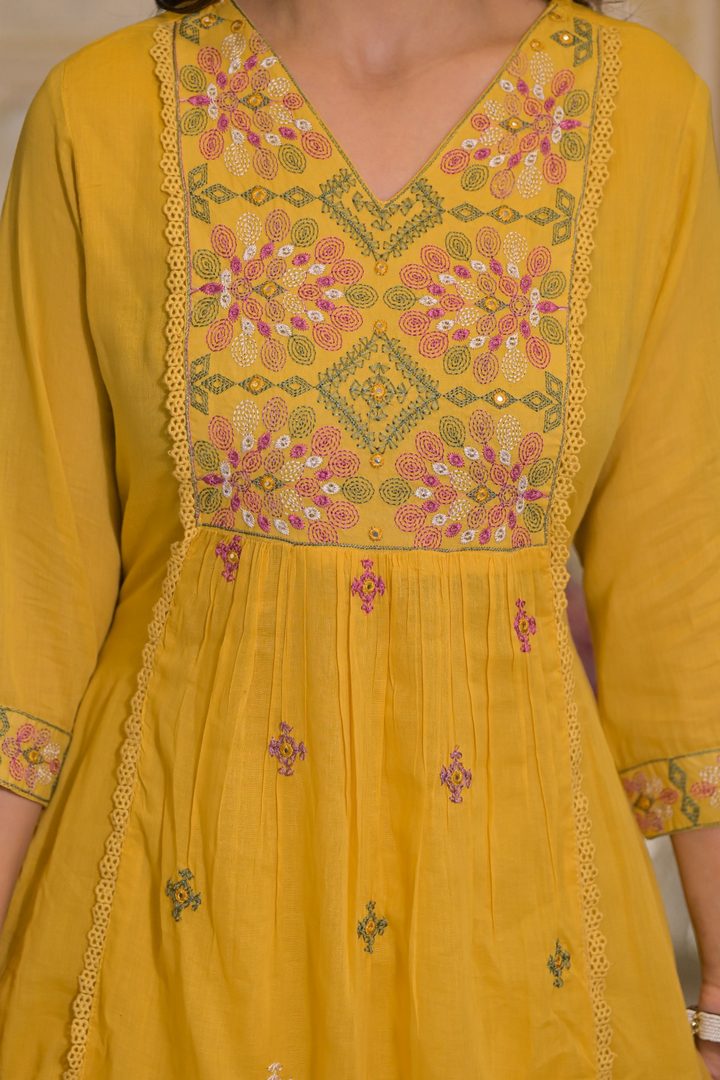 A close-up of a mustard yellow embroidered kurta featuring a V-neckline, a heavily decorated yoke with pink, green, and gold floral patterns, and matching embroidered cuffs.