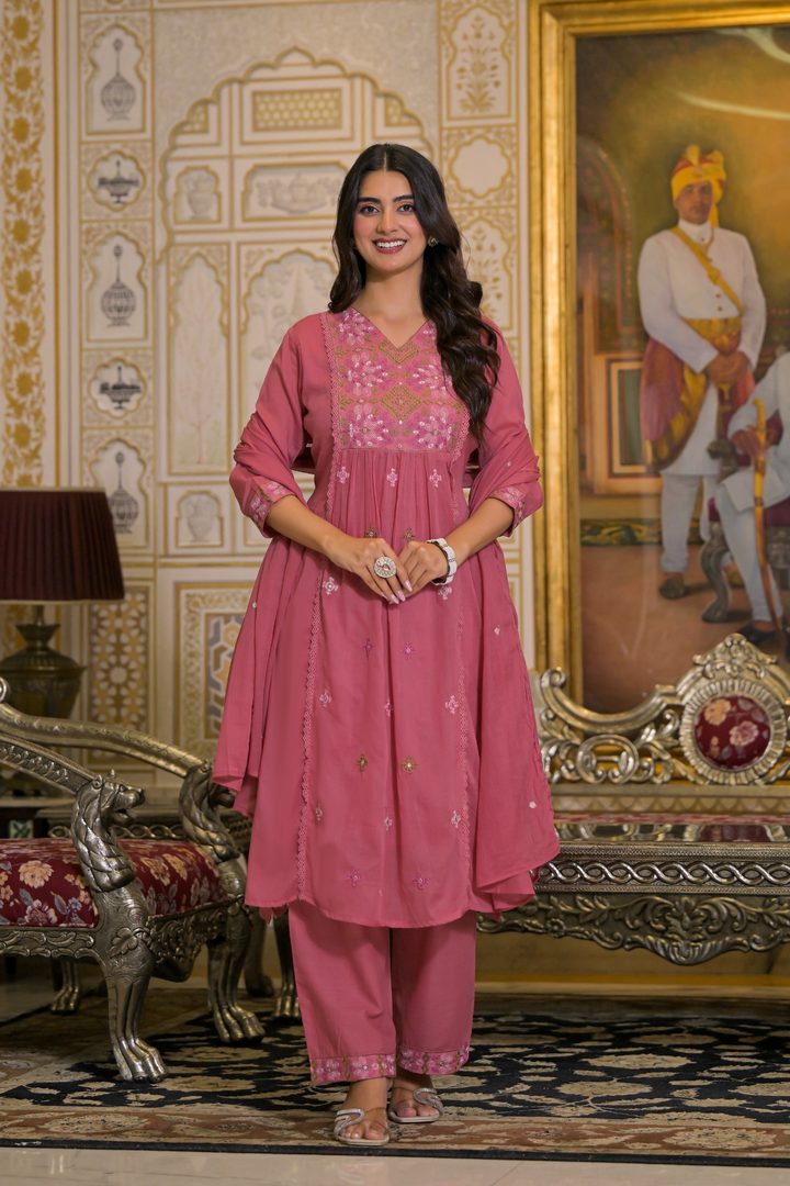 A woman models a pink traditional Indian salwar kameez suit with a matching dupatta in an ornate indoor setting. The outfit features a pink tunic with floral embroidery and matching trousers.