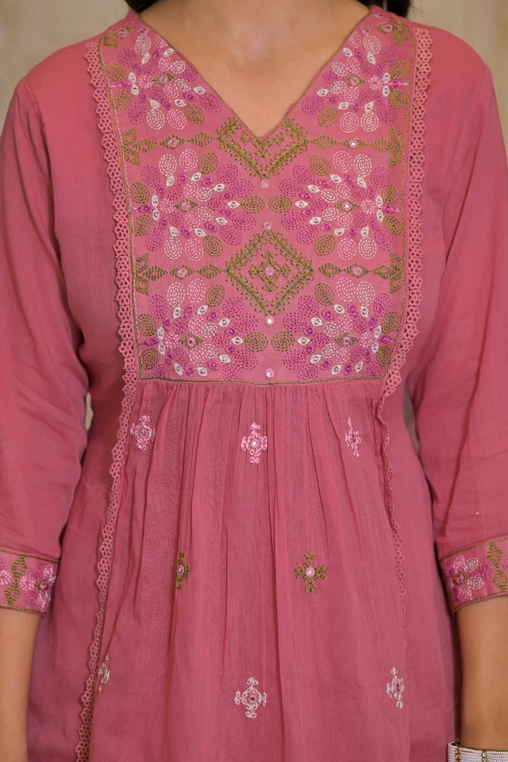 A close-up of a pink traditional embroidered tunic or kurta with a V-neckline, featuring intricate geometric and floral embroidery in olive green, white, and pink on the chest panel and scattered motifs on the skirt.