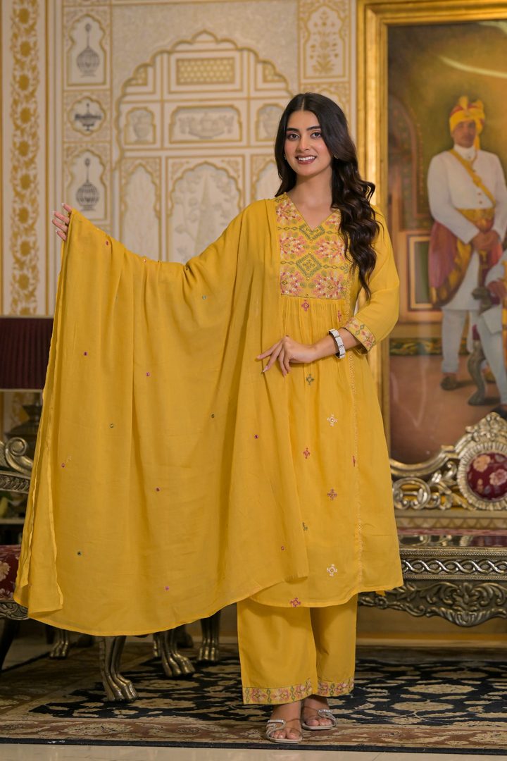 A woman models a traditional Indian salwar kameez suit in mustard yellow, featuring an embroidered kurta, matching trousers, and a sheer dupatta, posed in an ornate interior setting.