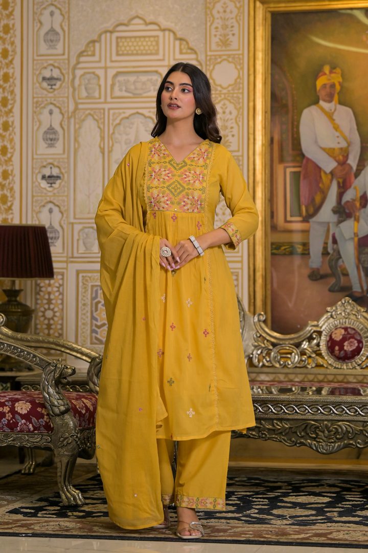A woman modeling a traditional Indian salwar kameez suit in mustard yellow with colorful floral embroidery, standing in an ornate, vintage-style room with gold-toned furniture and paintings.