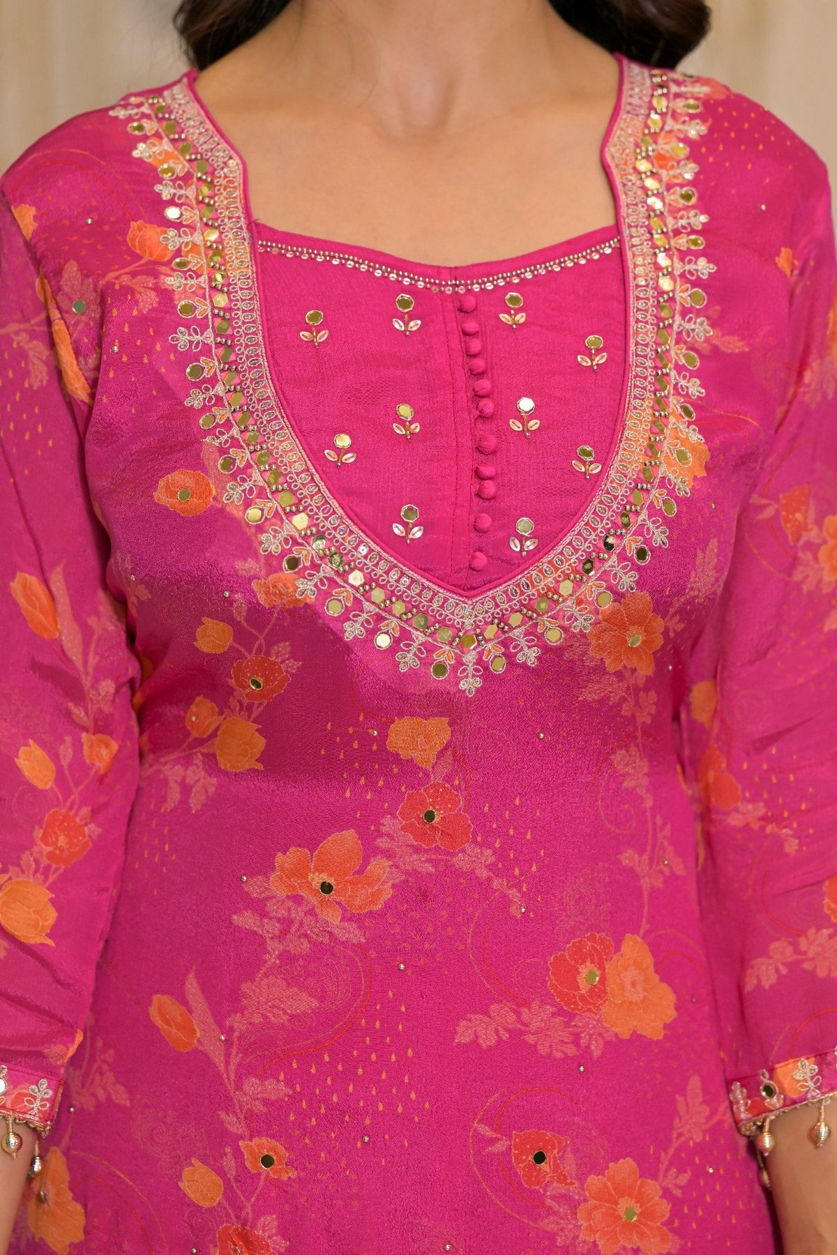 A close-up of a bright pink traditional tunic with orange floral patterns and ornate gold and silver mirror work embroidery around the neckline.