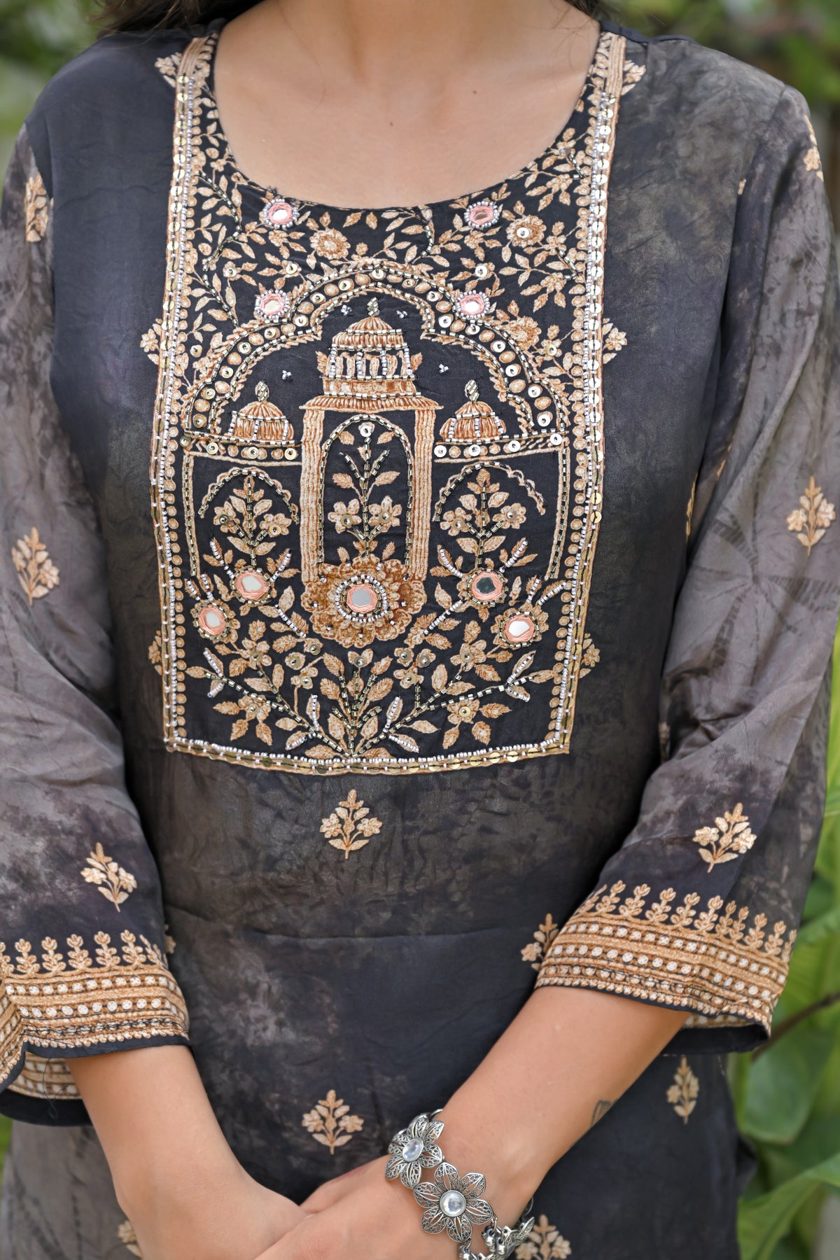 A close-up of a woman wearing a dark gray embroidered kurta with intricate gold, beige, and pink floral and arch motifs on the chest and cuffs.