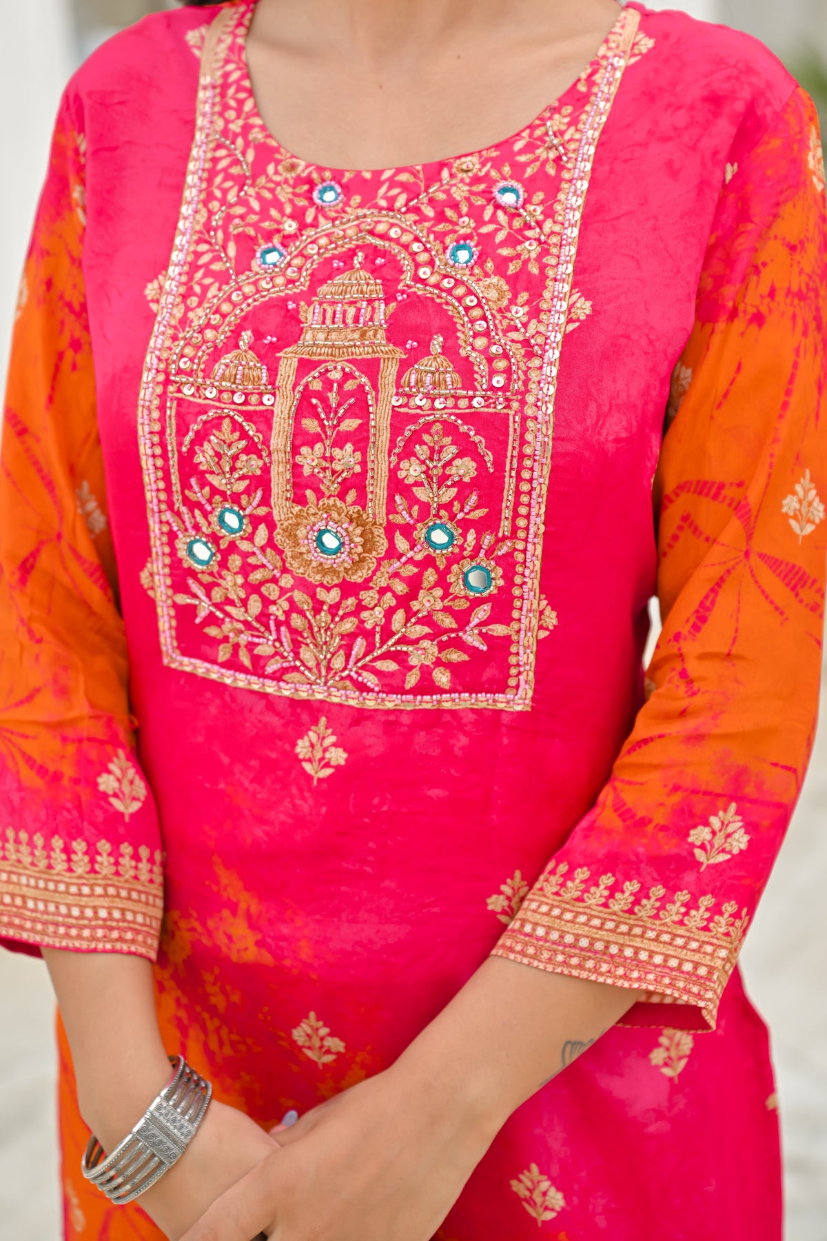 A close-up of a woman wearing a bright pink and orange embroidered kurta with gold detailing and turquoise accents on the neckline.