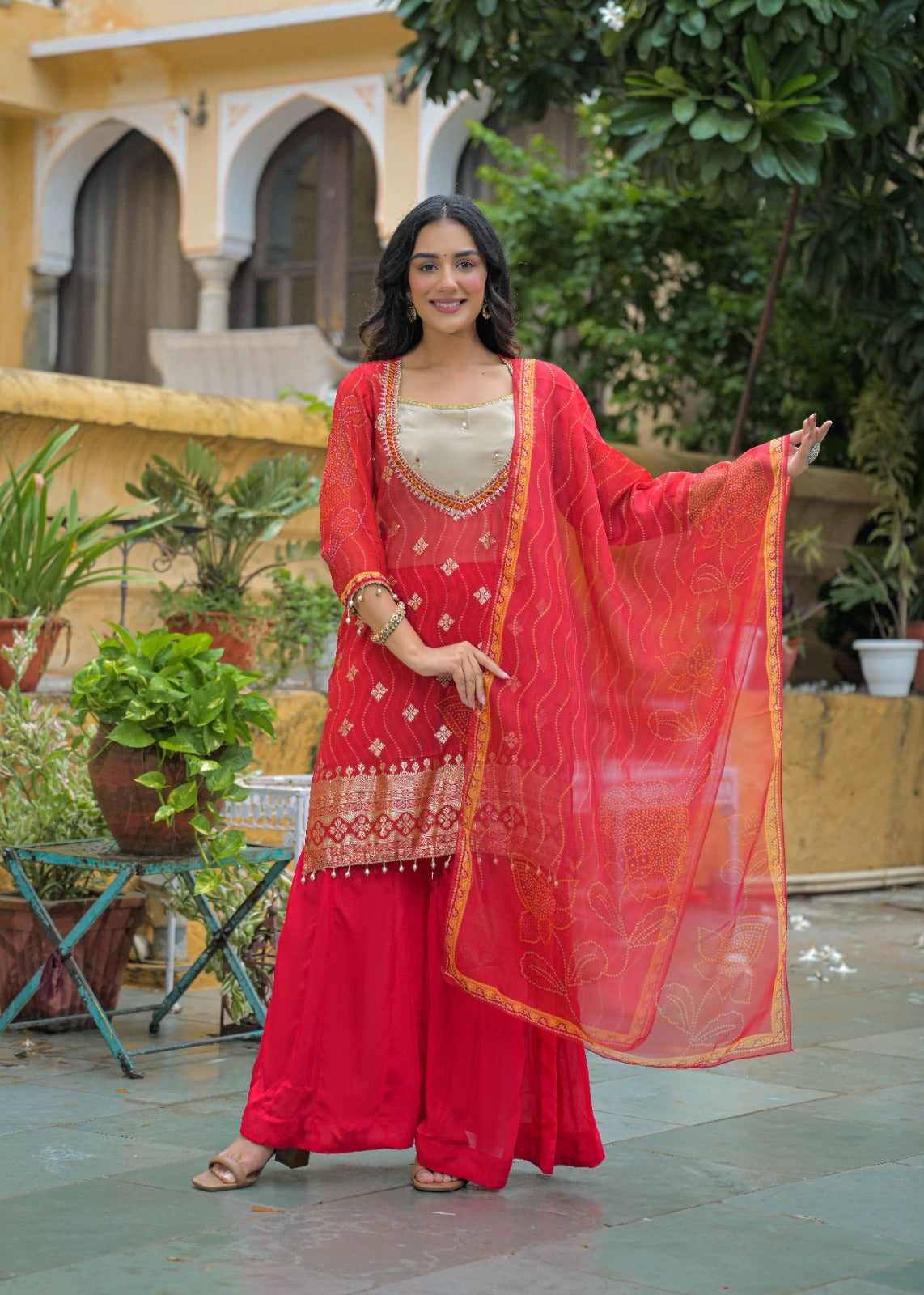 A woman models a traditional Indian red sharara suit with a beige embroidered blouse and a sheer red dupatta, set against a courtyard background with plants and a yellow building.