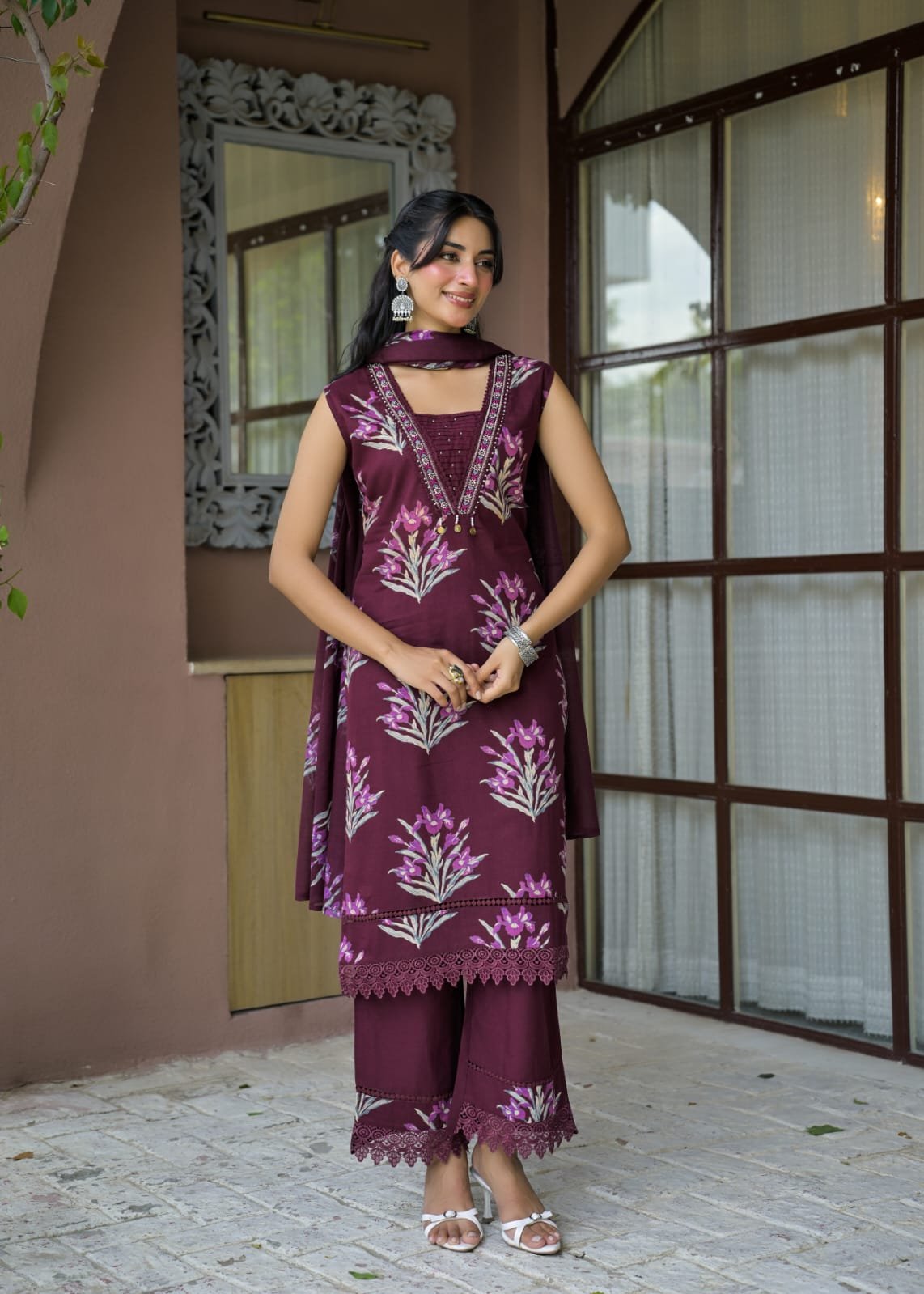 A woman models a three-piece traditional Indian salwar kameez suit in deep maroon with light pink and white floral embroidery. The set includes a sleeveless kameez top, matching trousers, and a dupatta, styled with silver jewelry against a rustic brown stucco wall background.