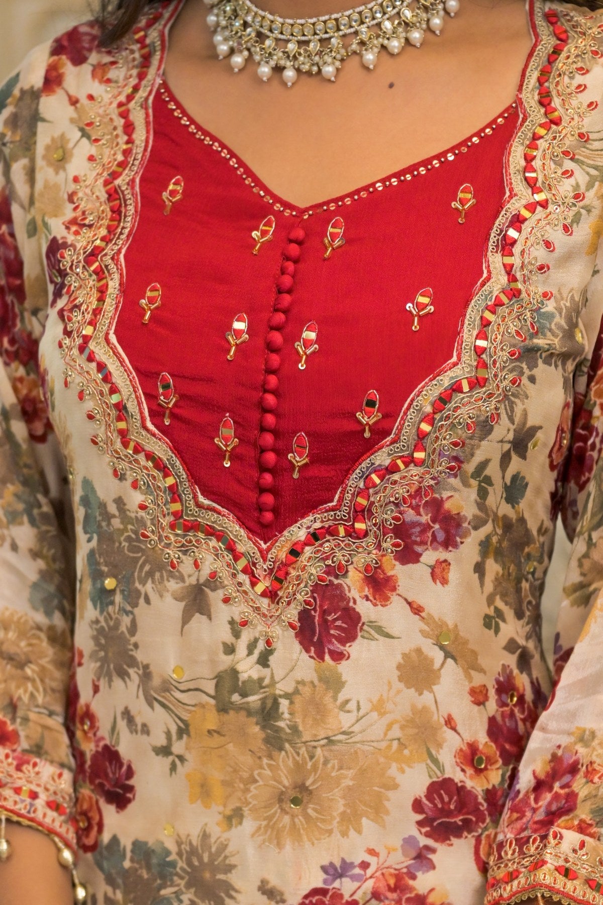 A close-up of a traditional embroidered kurta or tunic with a beige floral base, a bright red V-neck panel, and ornate gold and multicolor embroidery with scalloped borders.
